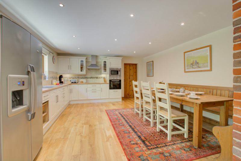 White fronted cupboards, stainless steel cooker and hob. Large dining table on rug on wooden floor.